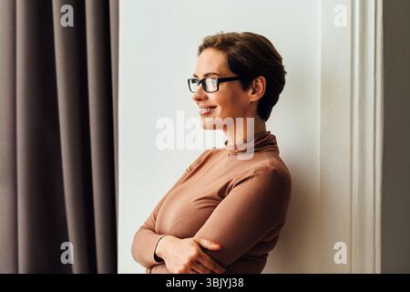 Side view of a beautiful businesswoman in eyeglasses standing in hotel room and looking at window Stock Photo