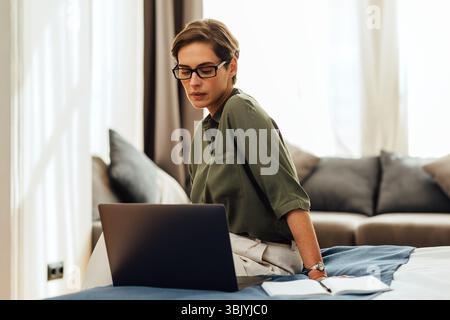 Caucasian woman in casual clothes sitting on bed in hotel room using laptop. Businesswoman working from a modern apartment. Stock Photo