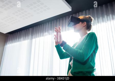 Woman wearing VR headset, smartwatch interacting with virtual interface in office by sheer curtains Stock Photo