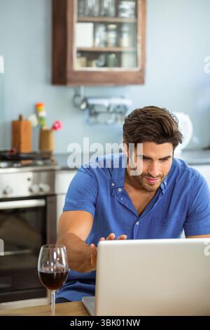 Silver laptop sitting on wooden table in kitchen near wine glass, spice rack, stainless steel stove Stock Photo
