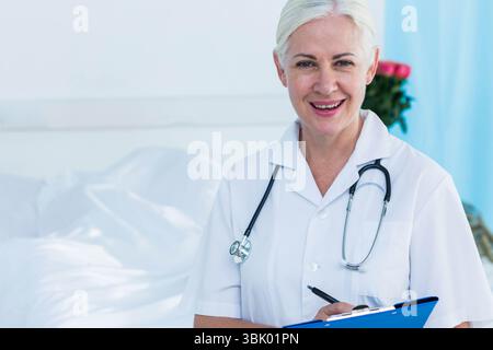 Doctor standing by bed in hospital room, holding blue clipboard and stethoscope near blue curtain Stock Photo