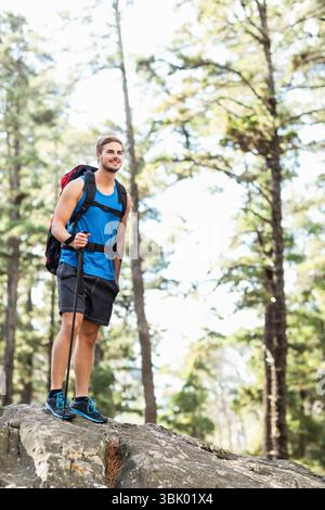 Male hiker standing atop rock outcrop in pine forest with backpack trekking pole and trail shoes Stock Photo