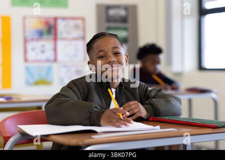 Smiling african american boy writing in notebook at school desk, enjoying classroom activities Stock Photo
