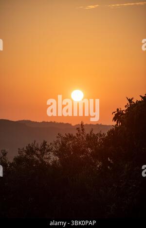 Fiery orb descends over rolling hills, casting a warm, golden hue. Nature's splendor unfolds. Stock Photo