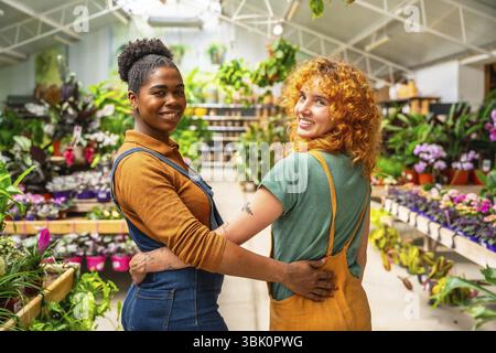 Two happy florists are hugging each other, smiling, while working in their plant nursery greenhouse Stock Photo