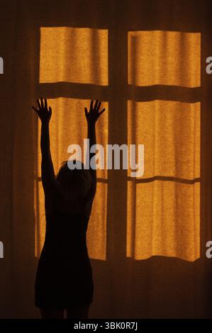 Back view of a woman with raised hands standing in front of a window with a red curtain Stock Photo
