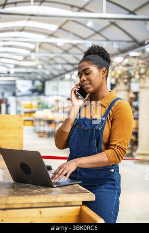 Confident black female employee managing orders and assisting customers in a garden center, using laptop and smartphone simultaneously Stock Photo