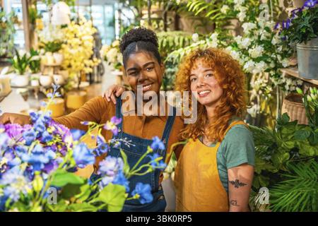 Two happy multiethnic female florists are smiling and posing surrounded by colorful flowers and plants in their flower shop Stock Photo