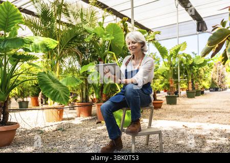 Senior woman working in plant nursery using digital tablet, managing plants and inventory in a greenhouse Stock Photo