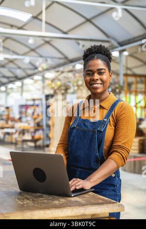 Confident young woman managing greenhouse inventory using a laptop, showcasing modern agriculture practices Stock Photo