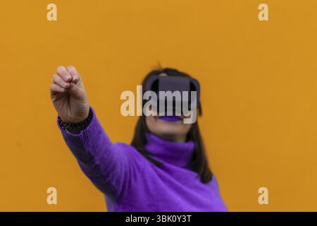 Portrait of a smiling woman wearing virtual reality goggles with the gesture of picking up something imaginary with her hand on a yellow background. s Stock Photo