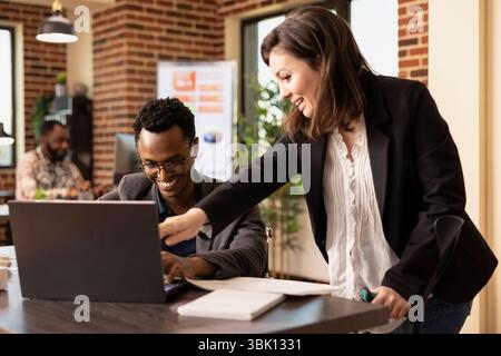 Black man sharing a laughter with his caucasian female coworker at the office desk while having a business discussion. Cheerful multiethnic colleagues having a meeting, reviewing reports on laptop. Stock Photo