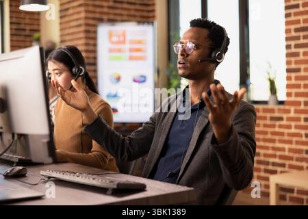 Male telemarketer raising hands in surprise and looking at computer screen during client call. Shocked customer support agent handling technical inquiries, frustrated by multiple incoming requests. Stock Photo