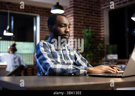 Male financial analyst works late at a brick wall office desk, reviewing documents and stock data on his laptop. The night setting creates a focused workspace for analyzing financial trends. Stock Photo