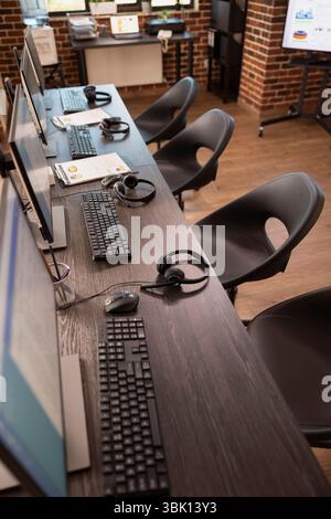 Vacant collaboration space in modern office, featuring multiple desks, digital monitors and headsets, ideal for customer support team. Empty table neatly organized with devices for group of employees. Stock Photo