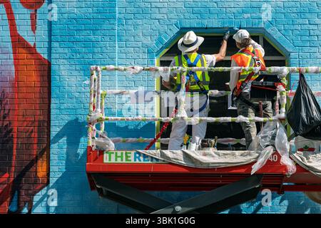 Two artists painting a colorful mural on a brick building while standing on a hydraulic lift. Downtown Calgary Alberta Canada Stock Photo