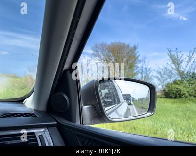 Driver side view captures a dusty car mirror reflecting a bus approaching from behind on a rural two lane road, framed by green and spring trees under Stock Photo