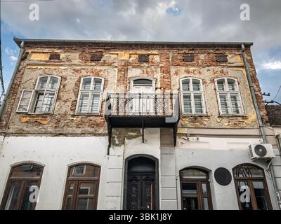 Two storey 19th century brick residence on central Valjevo street shows peeling plaster, rusted balcony and mismatched window repairs under a stormy s Stock Photo