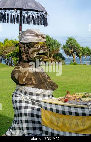 Bali, Indonesia - Dec 19, 2024: Statue and a table with a checkered tablecloth. The table is covered with food and a parasol is above it Stock Photo