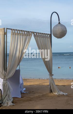 Beach scene with a white tent and white curtains for the wedding. The tent is set up on the beach and has a table and chairs. The beach is calm and pe Stock Photo