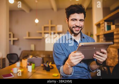 Bearded man sitting on wooden desk holding tablet in modern office, with coffee mugs, sticky notes Stock Photo
