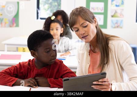 Female teacher using tablet to help african american boy in classroom, at school Stock Photo