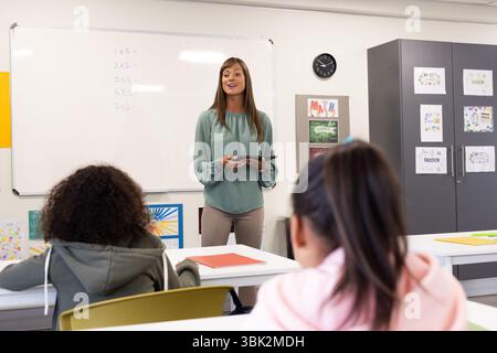 Female teacher engaging diverse students in classroom, holding tablet, discussing math, at school Stock Photo