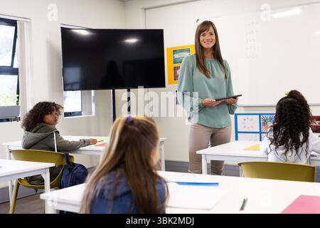 Female teacher holding tablet smiling at diverse students in classroom during lesson, at school Stock Photo