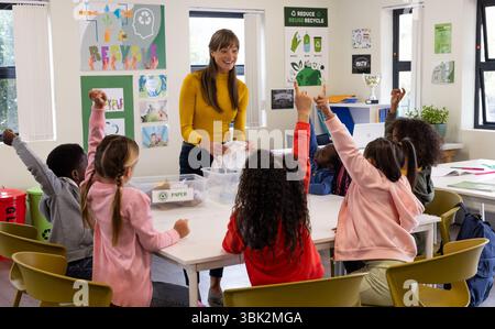 In school, female teacher engaging diverse students in recycling lesson, raising hands Stock Photo