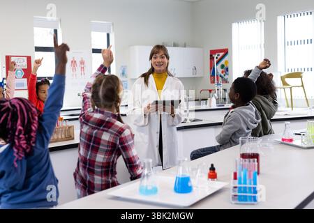 In school, female teacher engaging diverse students in science class, kids raising hands eagerly Stock Photo