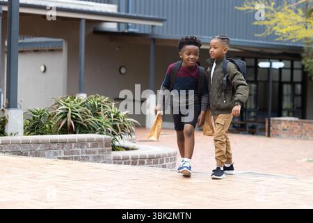 Two diverse boys walking outside school, carrying backpacks and lunch bags, smiling together Stock Photo
