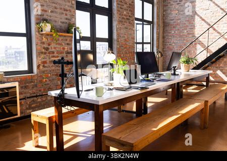 Modern office with computers and plants on long table, sunlight streaming in, copy space Stock Photo