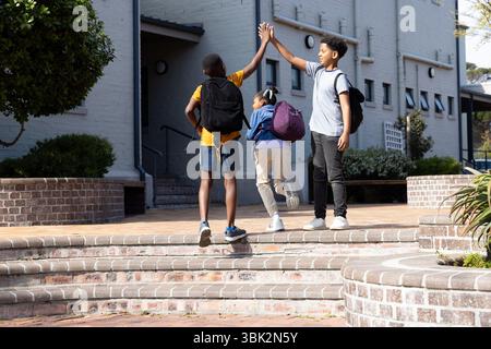 Outside school, diverse students giving high fives, wearing backpacks, excited Stock Photo