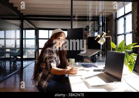 Woman coding at desk in modern office, writing notes and focusing on work Stock Photo