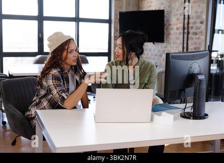 Diverse women collaborating on coding project using laptop and desktop in modern office Stock Photo