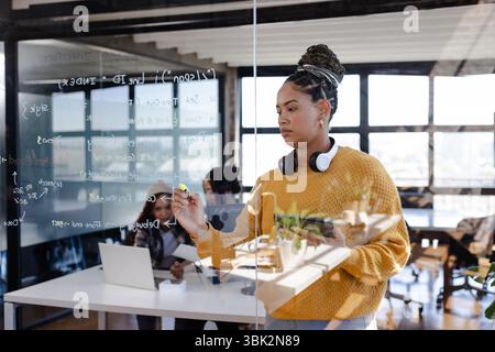 Confident woman writing code on glass board in modern office setting Stock Photo