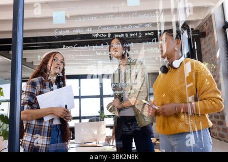 Diverse women collaborating on coding project, writing on glass board in modern office Stock Photo