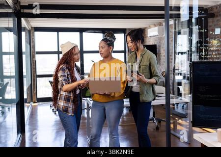 Diverse women collaborating on laptop and tablet in modern office, discussing coding project Stock Photo