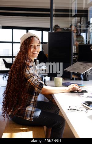 Smiling woman coding at desk with laptop and dual monitors in office Stock Photo