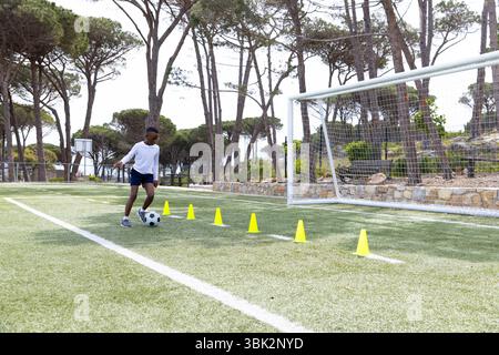 Practicing soccer, african american boy dribbling through cones on field, focused and determined Stock Photo