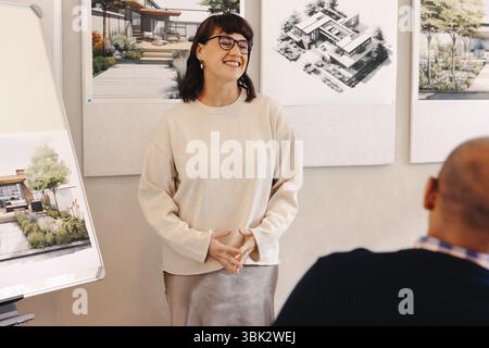 A woman confidently explains architectural designs in a professional setting. She stands near displayed design boards, showcasing innovations in archi Stock Photo