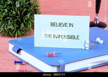 Motivational quote. BELIEVE IN THE IMPOSSIBLE text written on a card on a blue notebook next to colorful pins and an ornamental plant Stock Photo