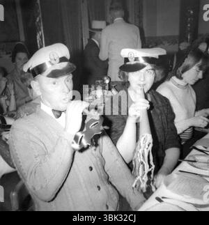 England. 1960s – A middle-aged couple are sitting at a dining table wearing sailor hats and blowing party horns. The man is wearing a bow tie and holding a mask. The woman is wearing cat-eye style spectacles. Stock Photo
