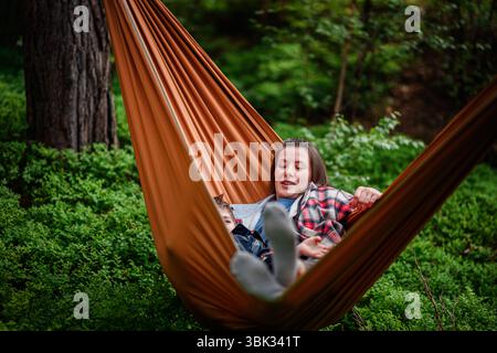 Smiling boy and relaxed woman lying in an orange hammock in a green forest, enjoying nature and quality time together. Stock Photo