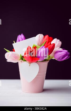 Pink bucket holding colorful tulips, blank white card and heart tag on table by purple backdrop Stock Photo