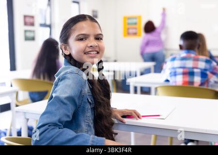 Smiling girl in classroom looking back, holding pencil, female teacher writing on board, at school Stock Photo