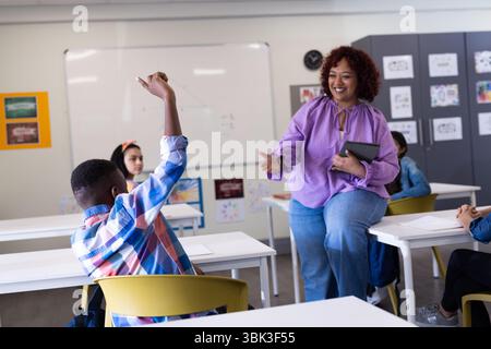 Female teacher engaging with diverse students in class, boy raising hand to participate, at school Stock Photo