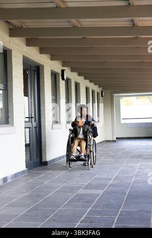 Two diverse boys in school hallway, one pushing other in wheelchair, smiling together Stock Photo