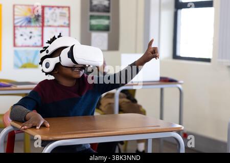 African american boy using vr headset in classroom, enthusiastically raising hand, at school Stock Photo