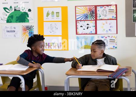 In school, two diverse boys sharing pencils and smiling while studying in classroom Stock Photo